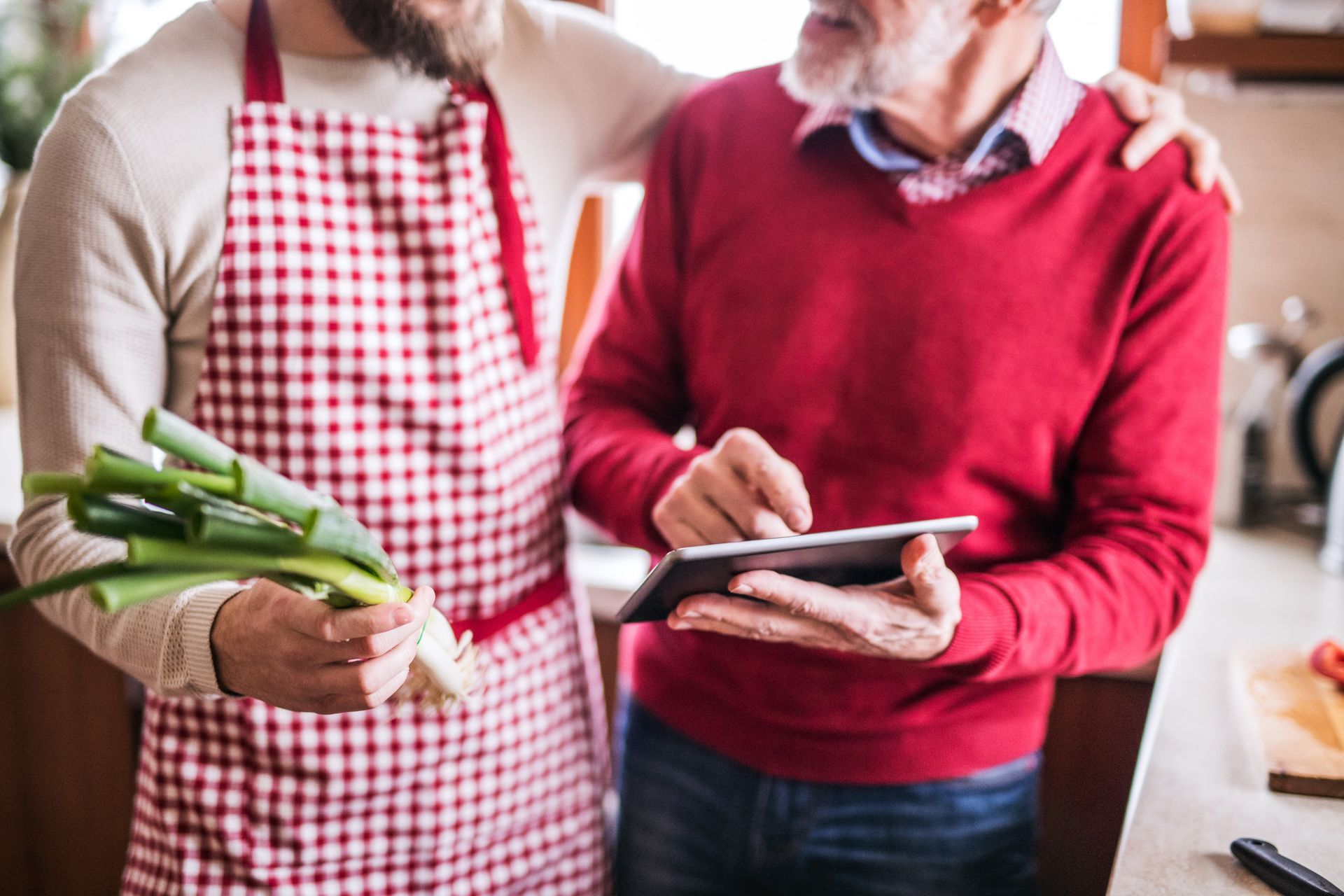 Two men cooking together in a kitchen. One holds green onions, the other looks at a tablet.