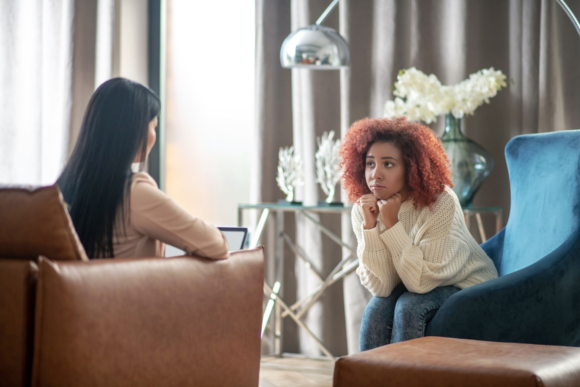 Two women in a therapy session; one speaks while the other looks distressed, hands clasped, in a blue armchair.