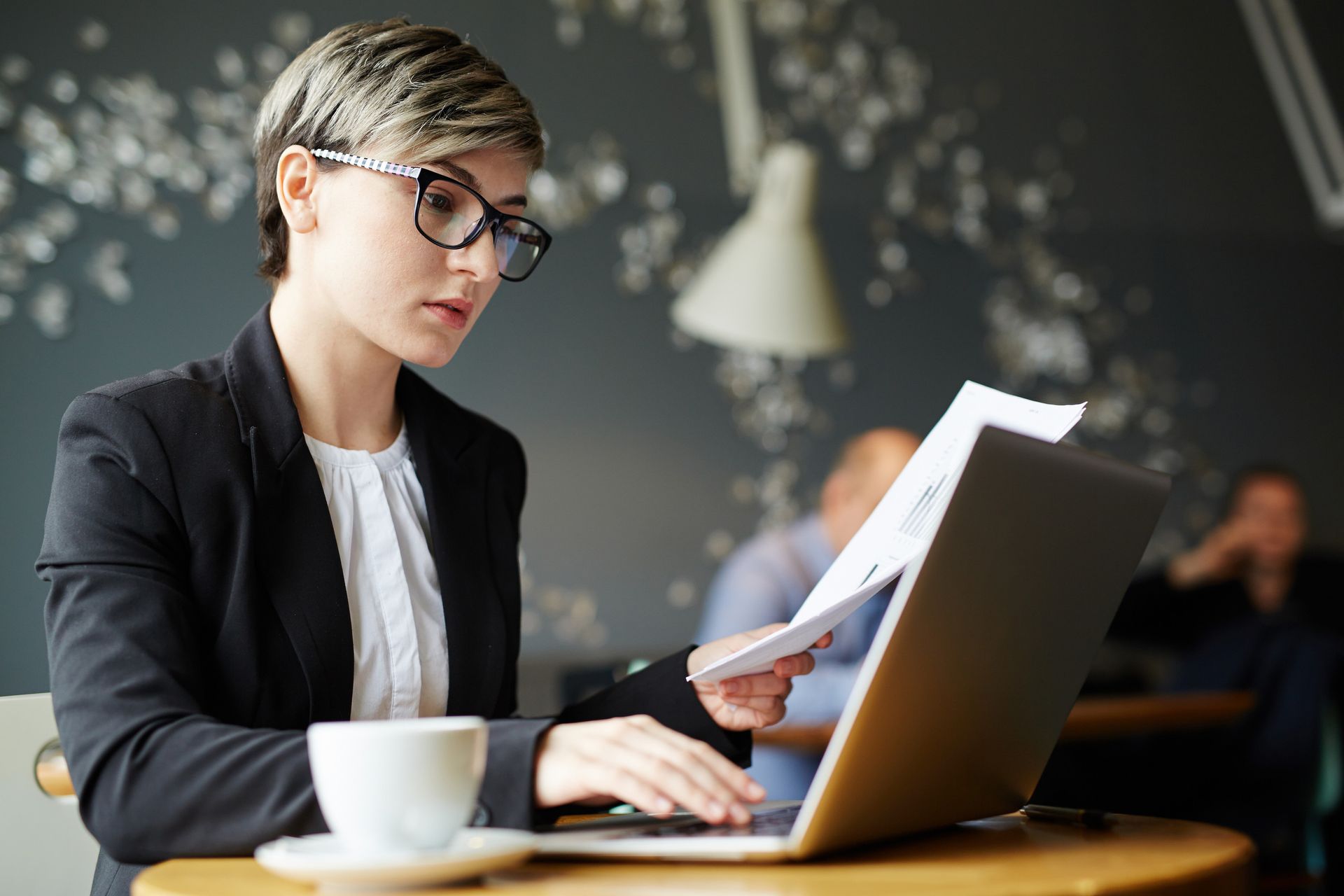 Woman in blazer at laptop, reading papers, cafe setting with coffee cup.