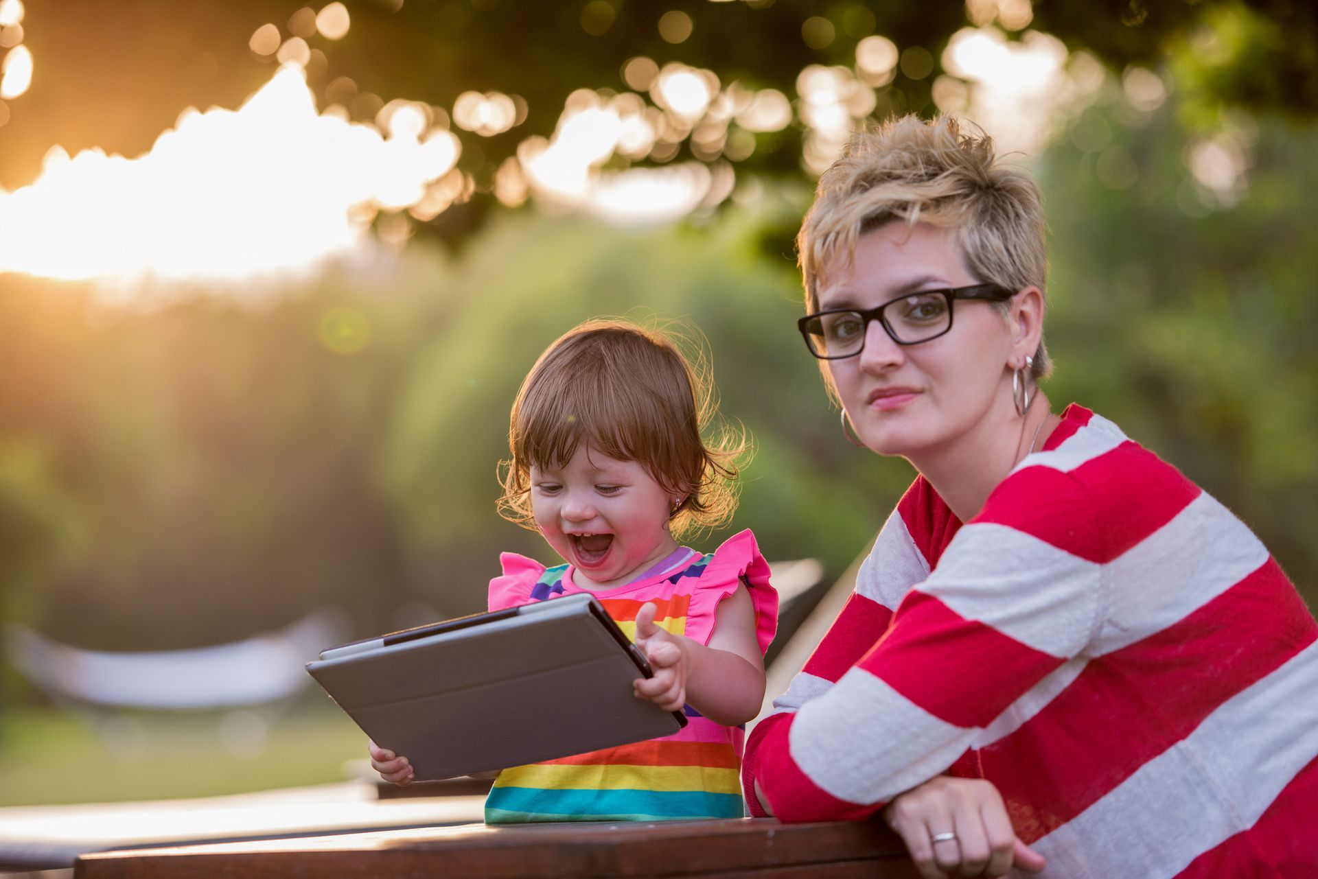 Woman with glasses smiles next to a laughing toddler, both using a tablet outside at sunset.