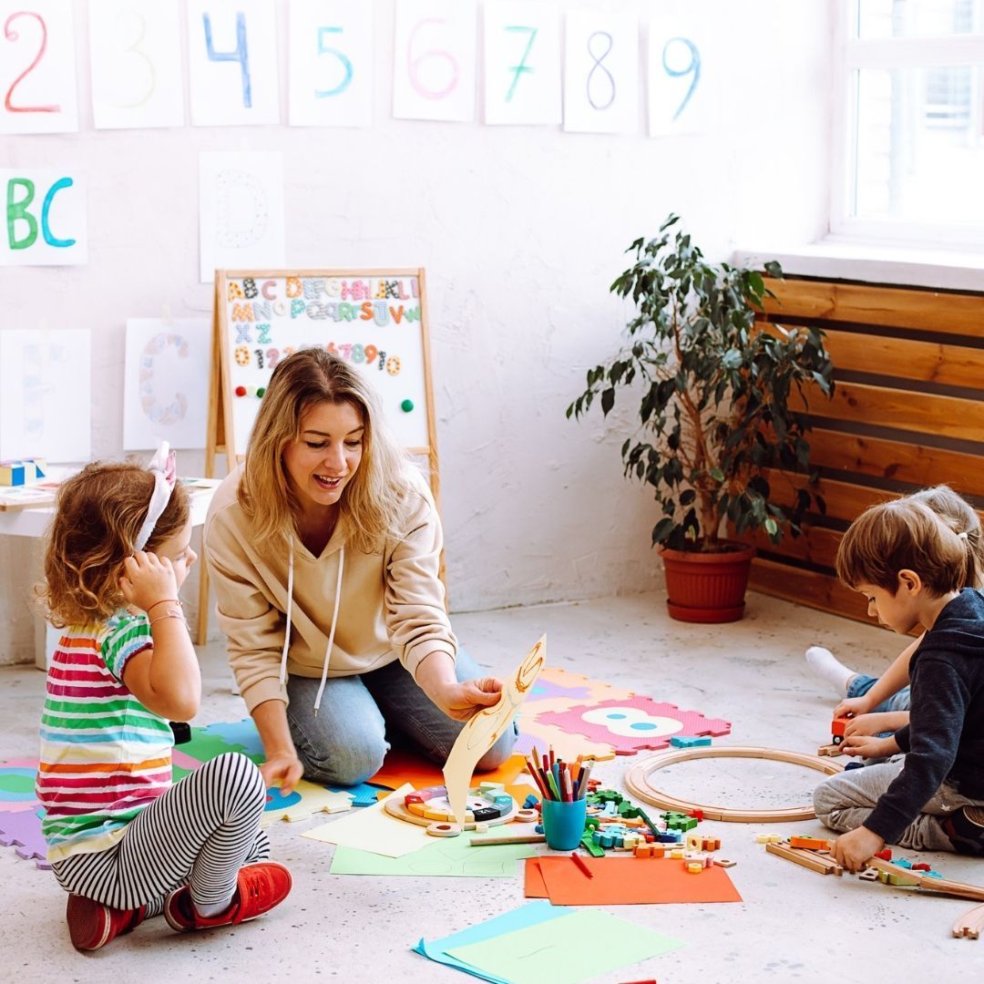 A teacher kneels with children on the floor, showing a wooden toy. Classroom setting, colorful toys.