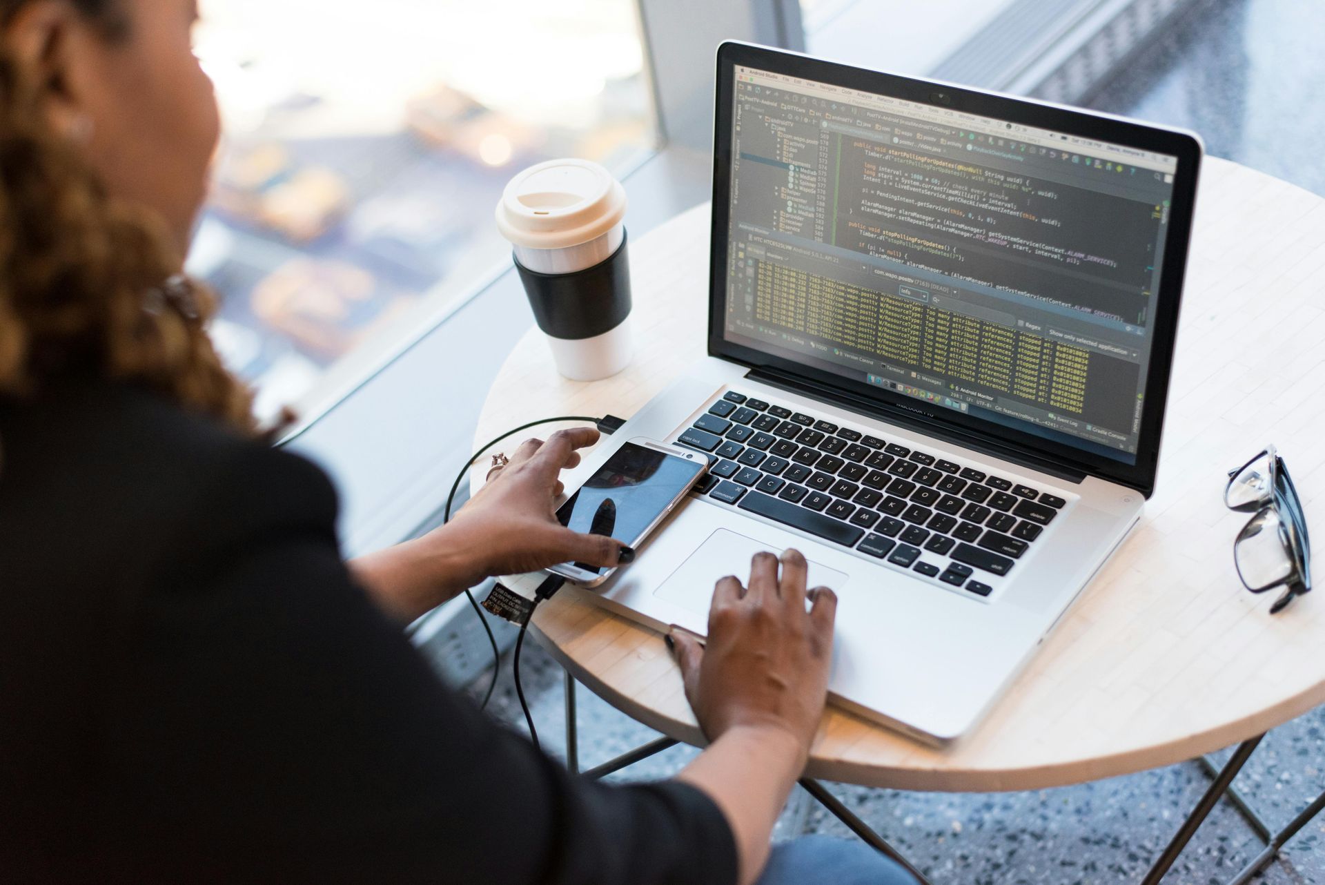Person coding on laptop, connected to a smartphone, with a cup of coffee and glasses on a table near a window.