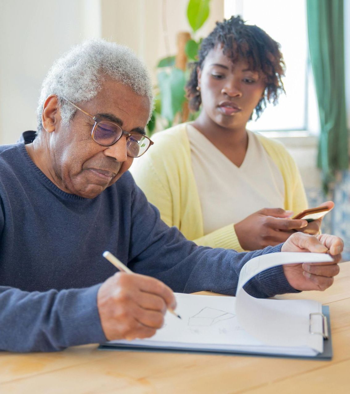 Elderly Black man in glasses writing on a clipboard, young Black woman looks on, holding phone.