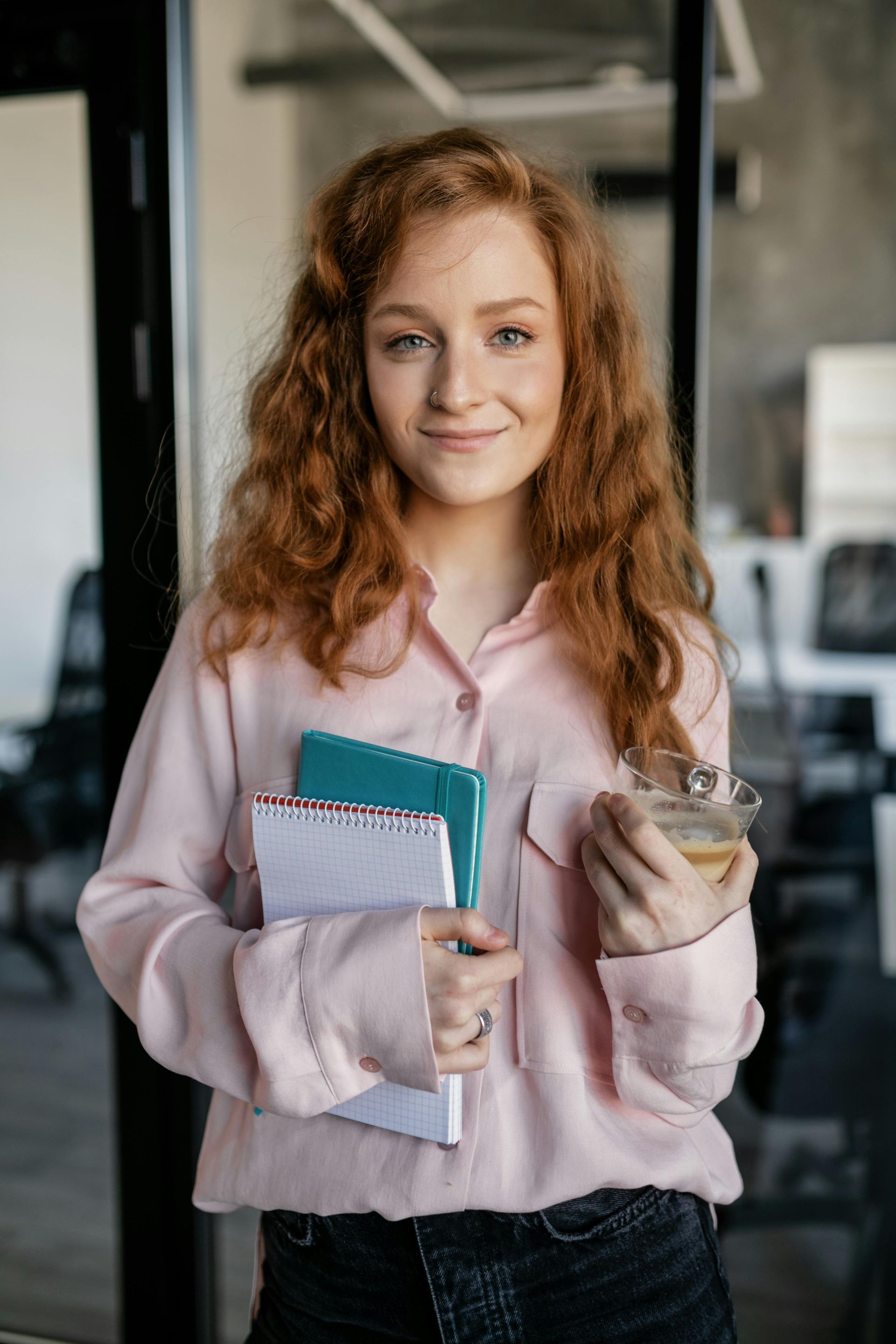 Woman with red curly hair in pink shirt holds notebooks and a cup, smiles in an office.