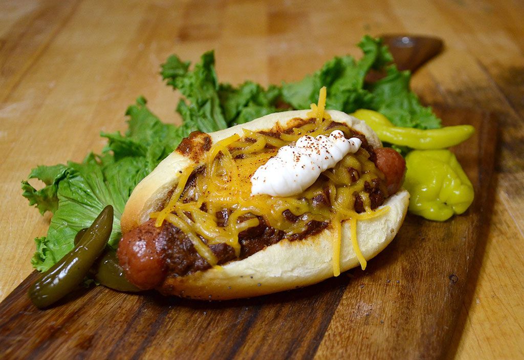 A chili dog on a bun with cheese and sour cream on a wooden cutting board.