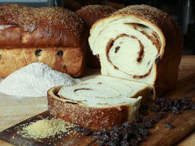 A loaf of bread is sitting on a wooden cutting board next to raisins and flour.