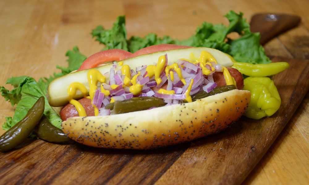 A hot dog with mustard , onions , pickles and lettuce on a wooden cutting board.