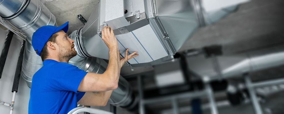 HVAC technician in blue uniform inspecting ventilation system ducts in a commercial setting.