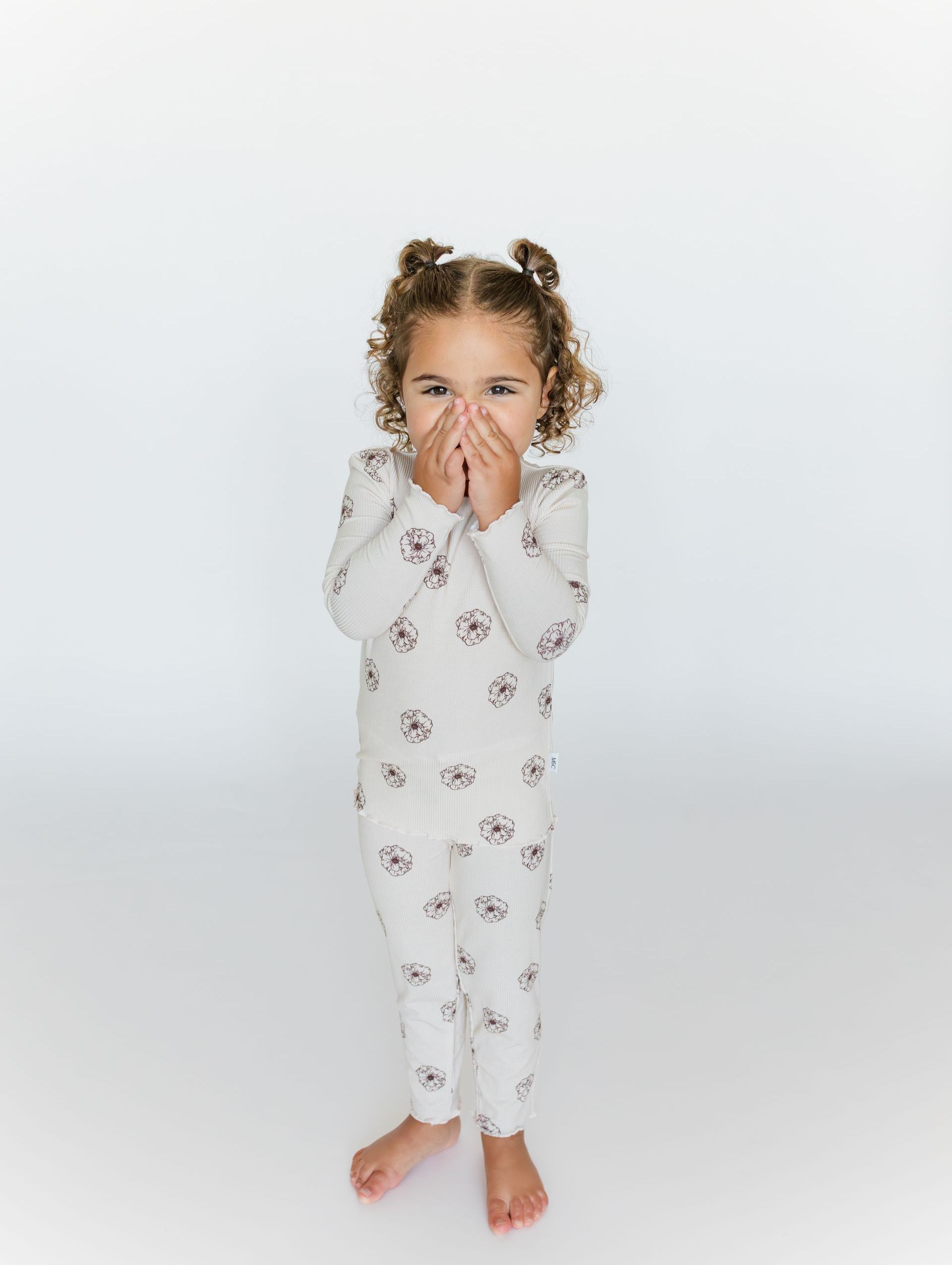Girl with curly brown hair in two buns, covering mouth, wearing a patterned Mica and Co two-piece pajama set. 