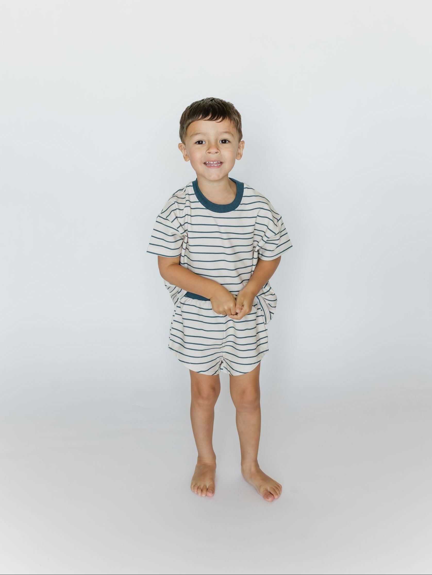 A young boy smiles while standing in a white room, wearing a matching striped top and shorts set.