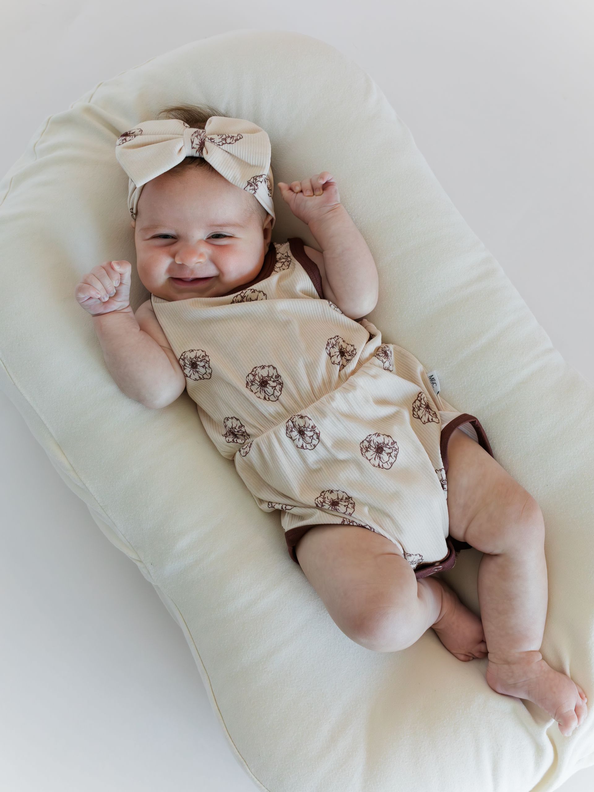 Baby in patterned romper and headband lying on a cream-colored cushioned surface, smiling.