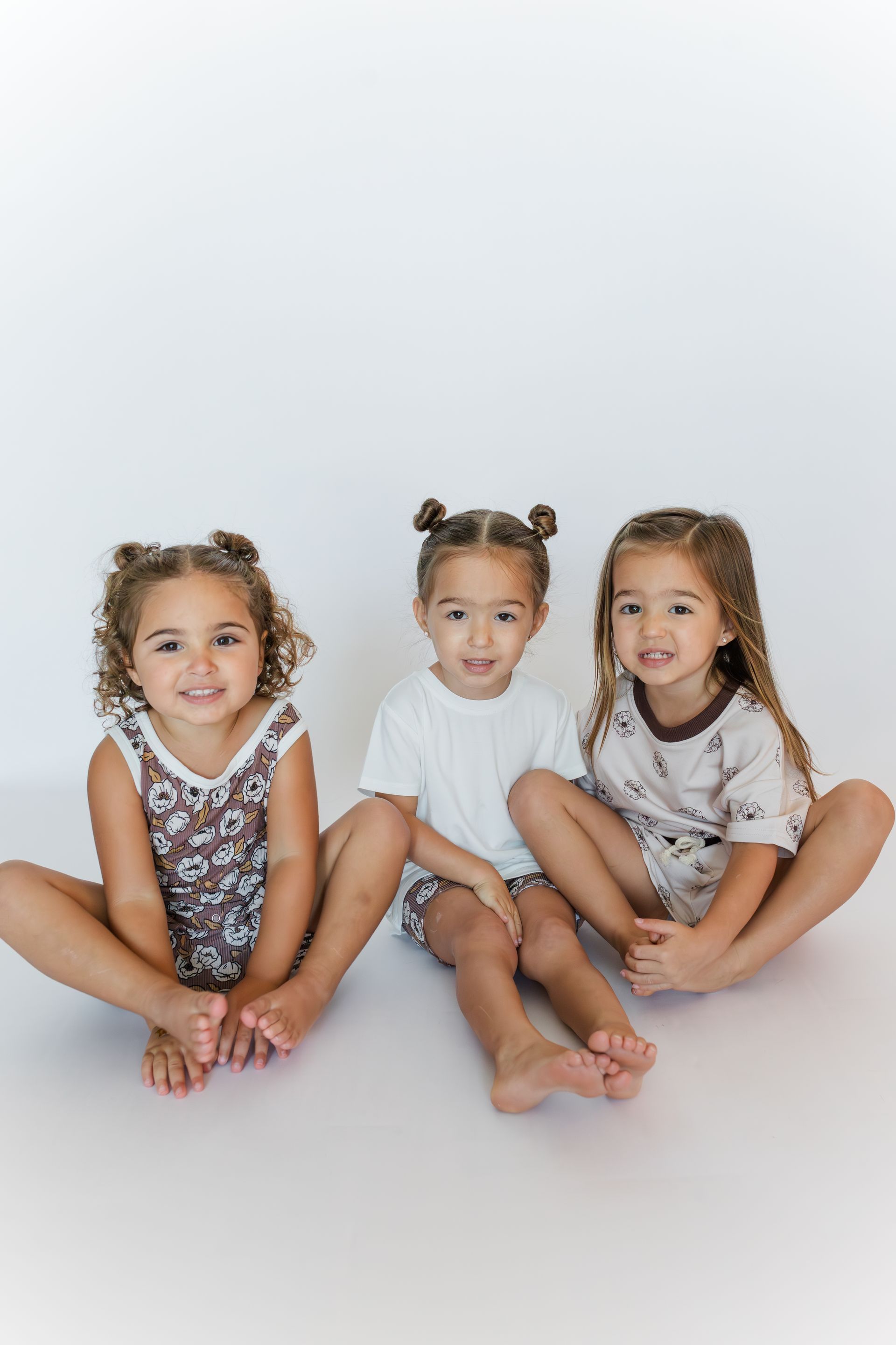 Three young girls sitting on the floor, smiling. They all have their hair up in buns and are wearing matching outfits.