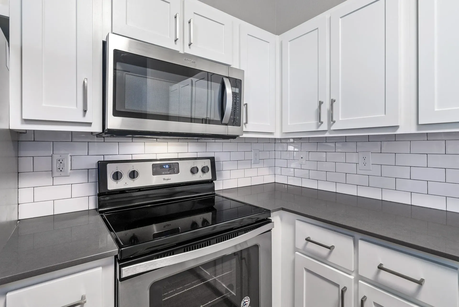 Bright kitchen with white cabinets, stainless steel microwave above a black electric range, gray countertops, and white subway tile backsplash.