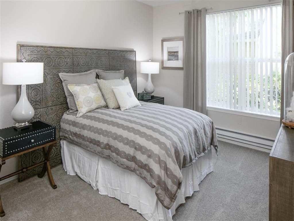 Bedroom in apartment with a large bed, nightstands, lamps, and a window with vertical blinds.