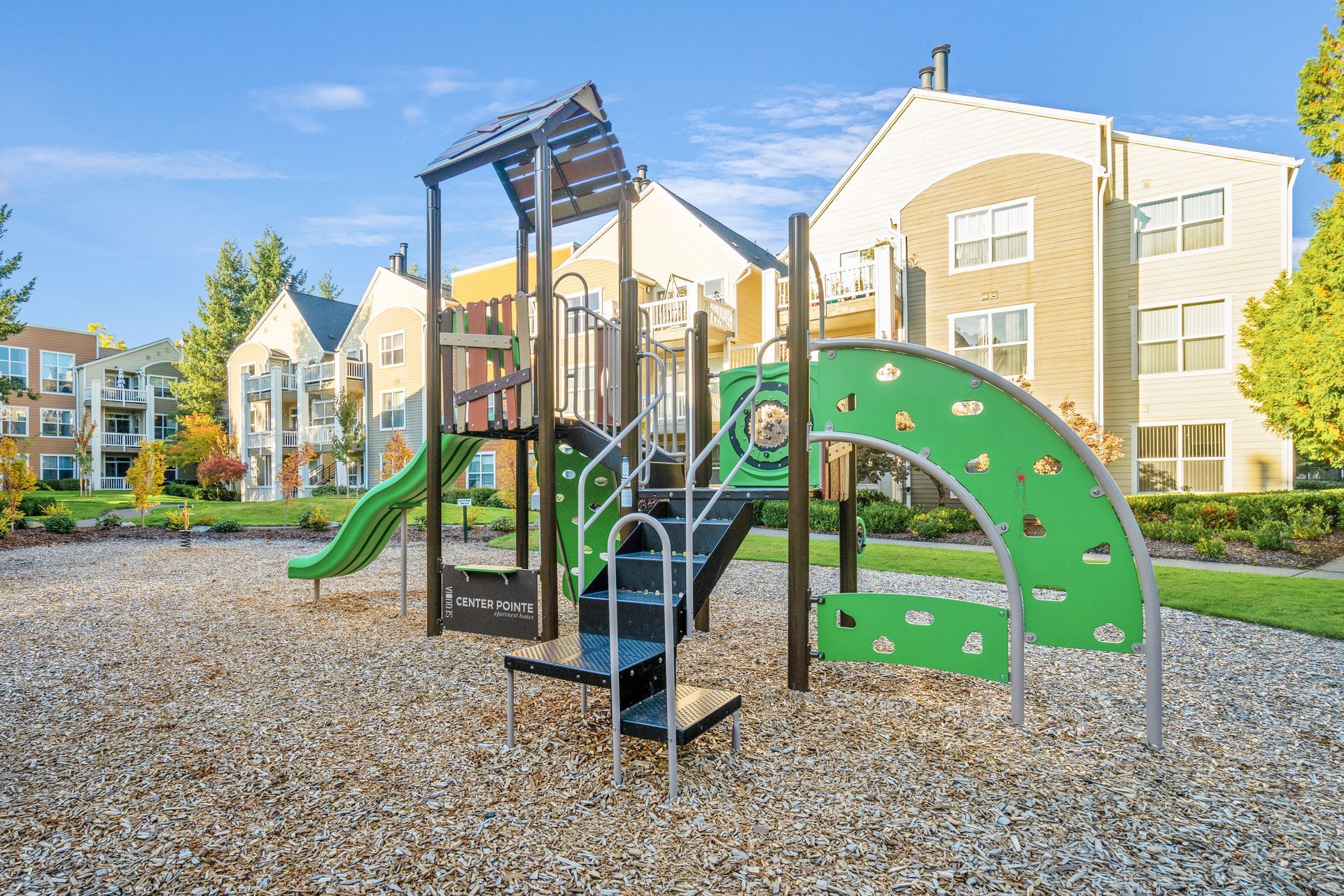 Playground with green slide, climbing wall, and stairs, in front of an apartment building.