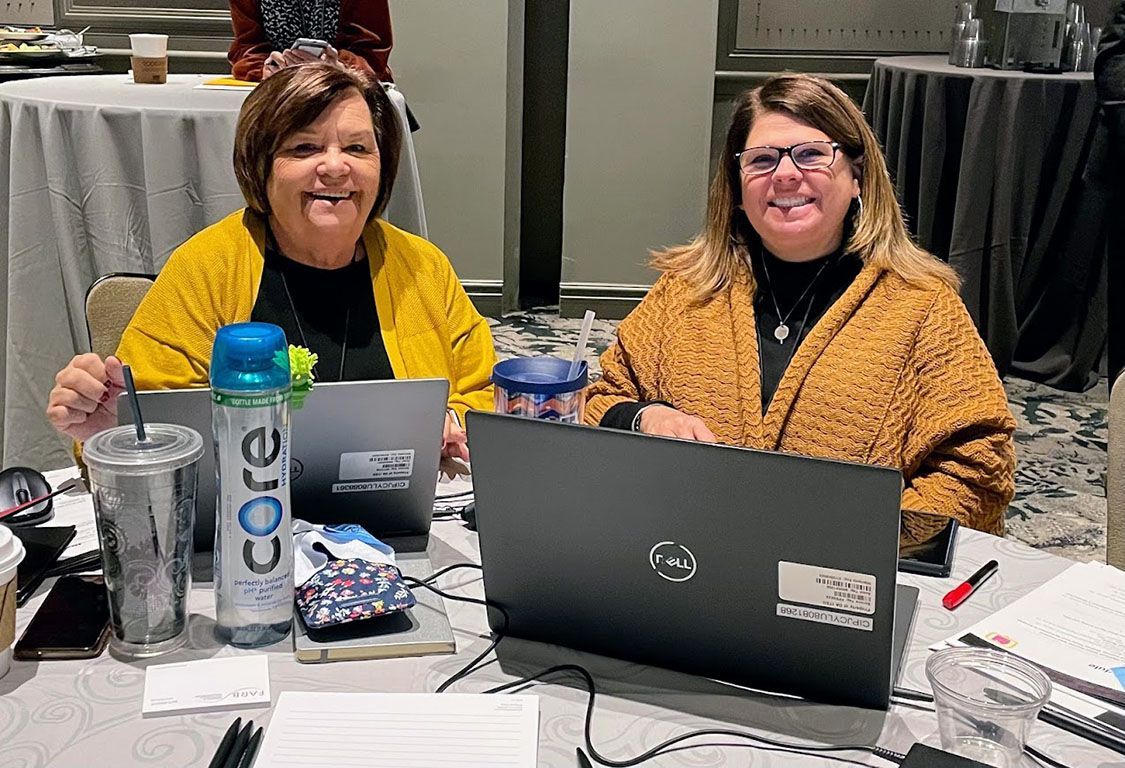 Two women are sitting at a table with laptops.