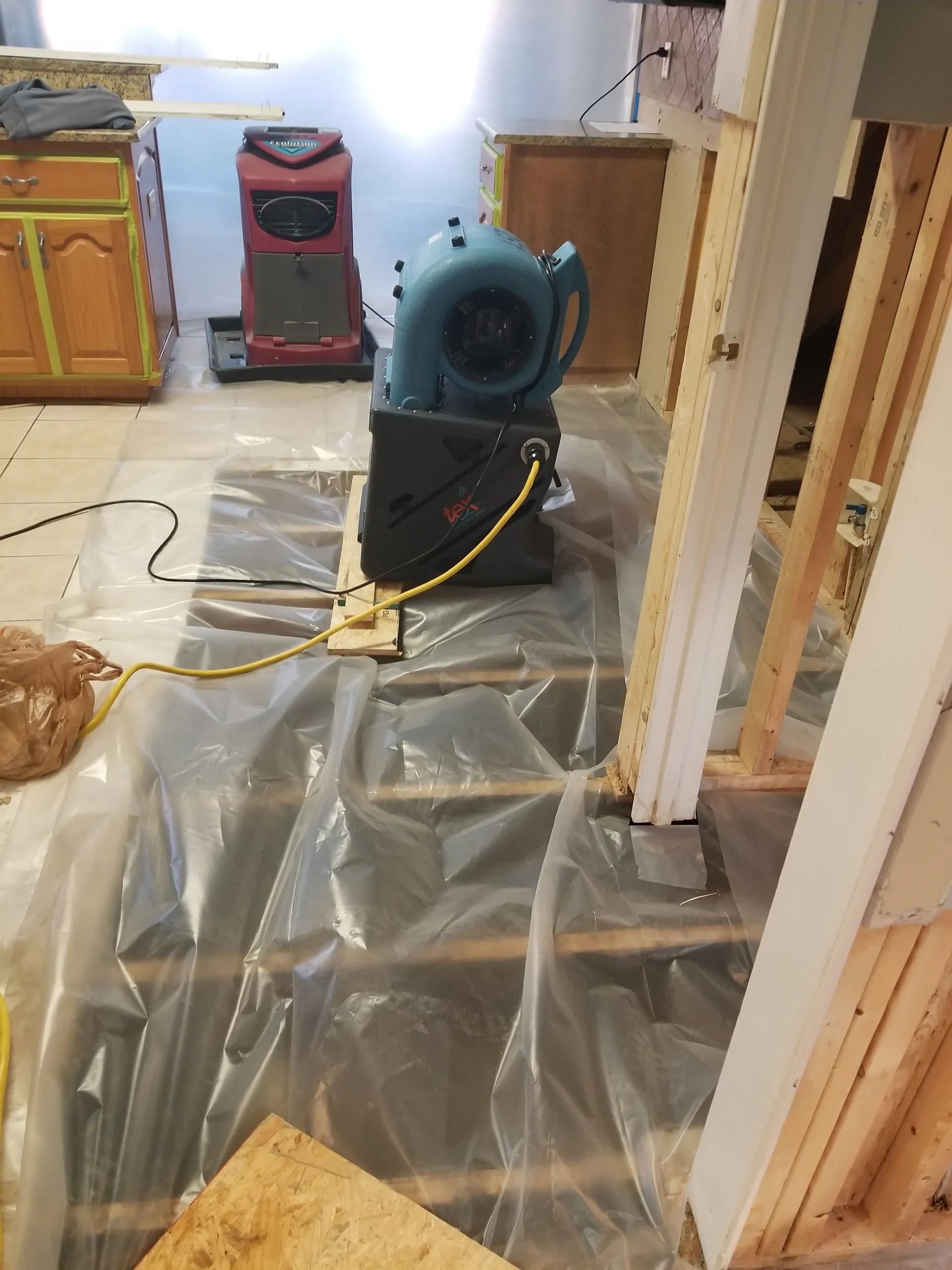 Large blue fan on plastic sheeting in a room under construction. Other equipment and exposed wood visible.