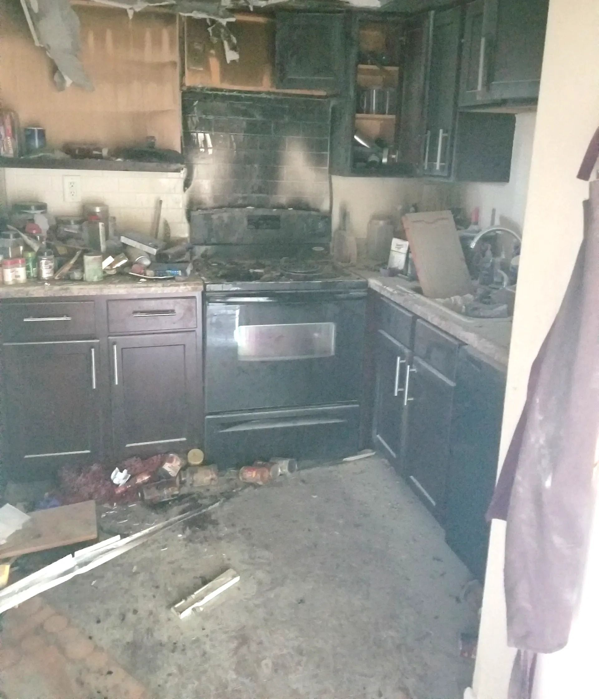 Kitchen heavily damaged by fire; cabinets, stove, and walls blackened with soot. Debris on the floor.