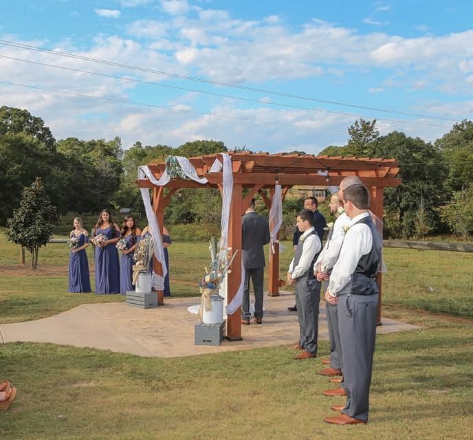Outdoor wedding ceremony under a wooden pergola, with bride, groom, attendants, and guests standing on the lawn