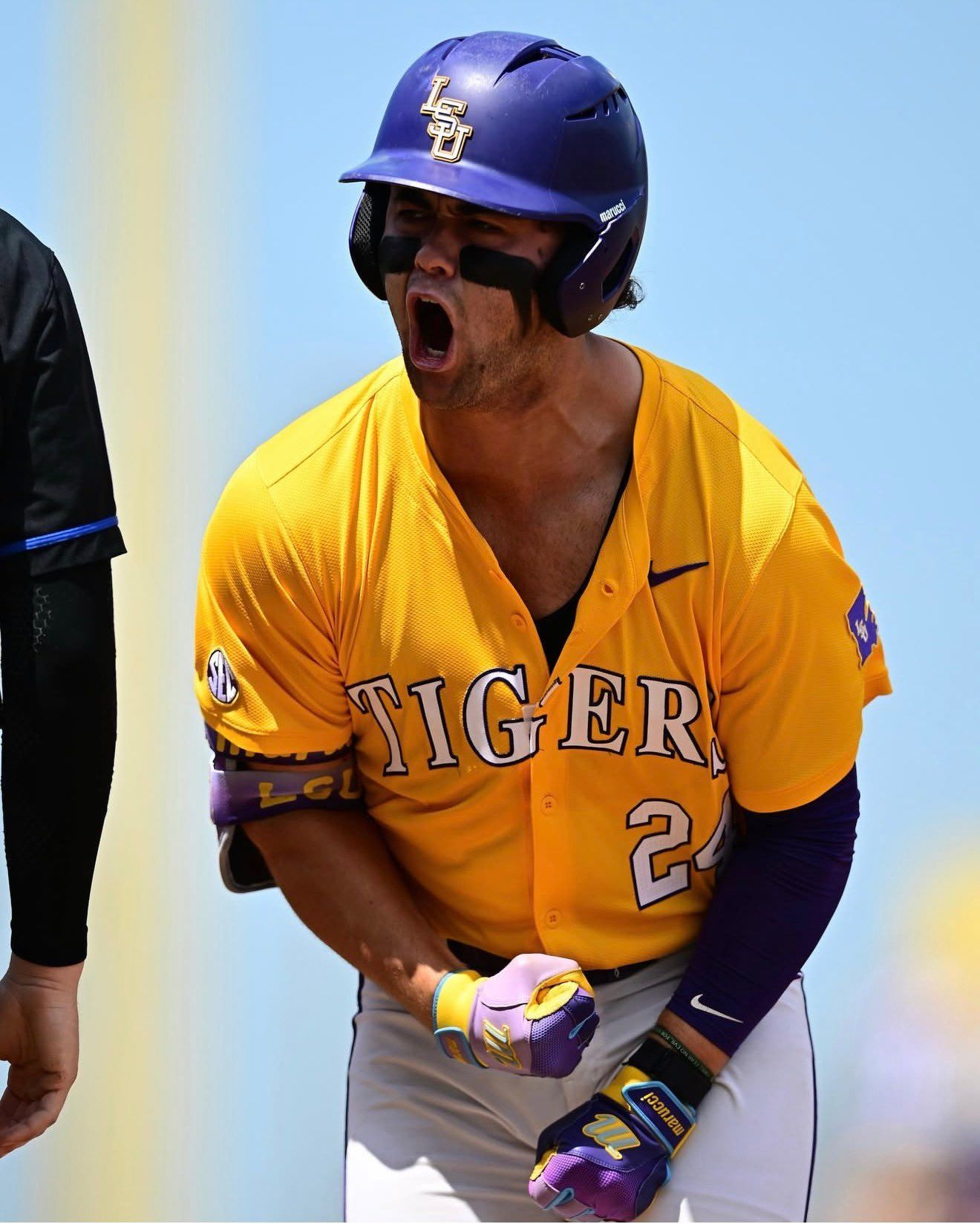 An LSU Tigers baseball player in a yellow jersey yells and pumps his fist in celebration while wearing a purple helmet.