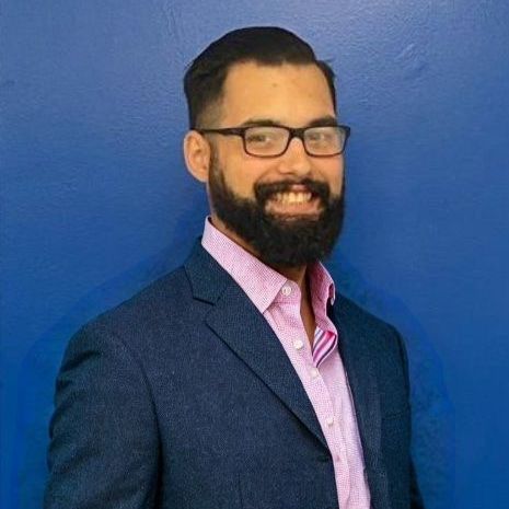 A man with a beard wearing glasses and a suit is standing in front of a blue wall.