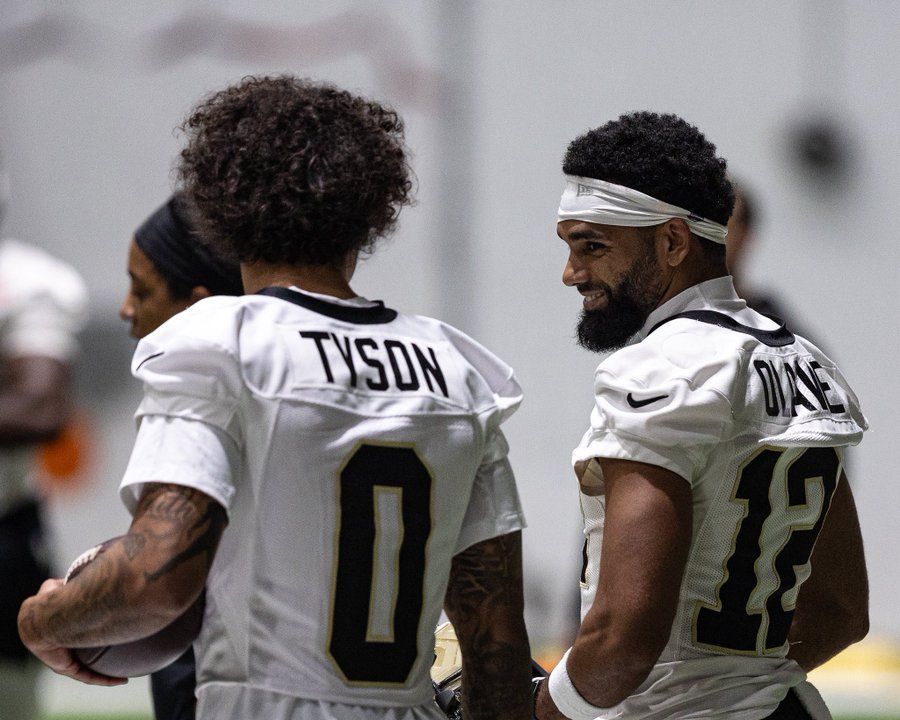 Two football players in white uniforms standing on the field, one with “TYSON” on his jersey.