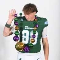 Football player in a green uniform wearing large purple and gold beads, holding up a hand