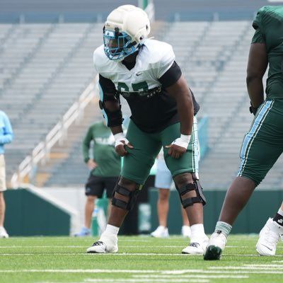 Football player in white and green crouches on the field during practice, with teammates nearby.