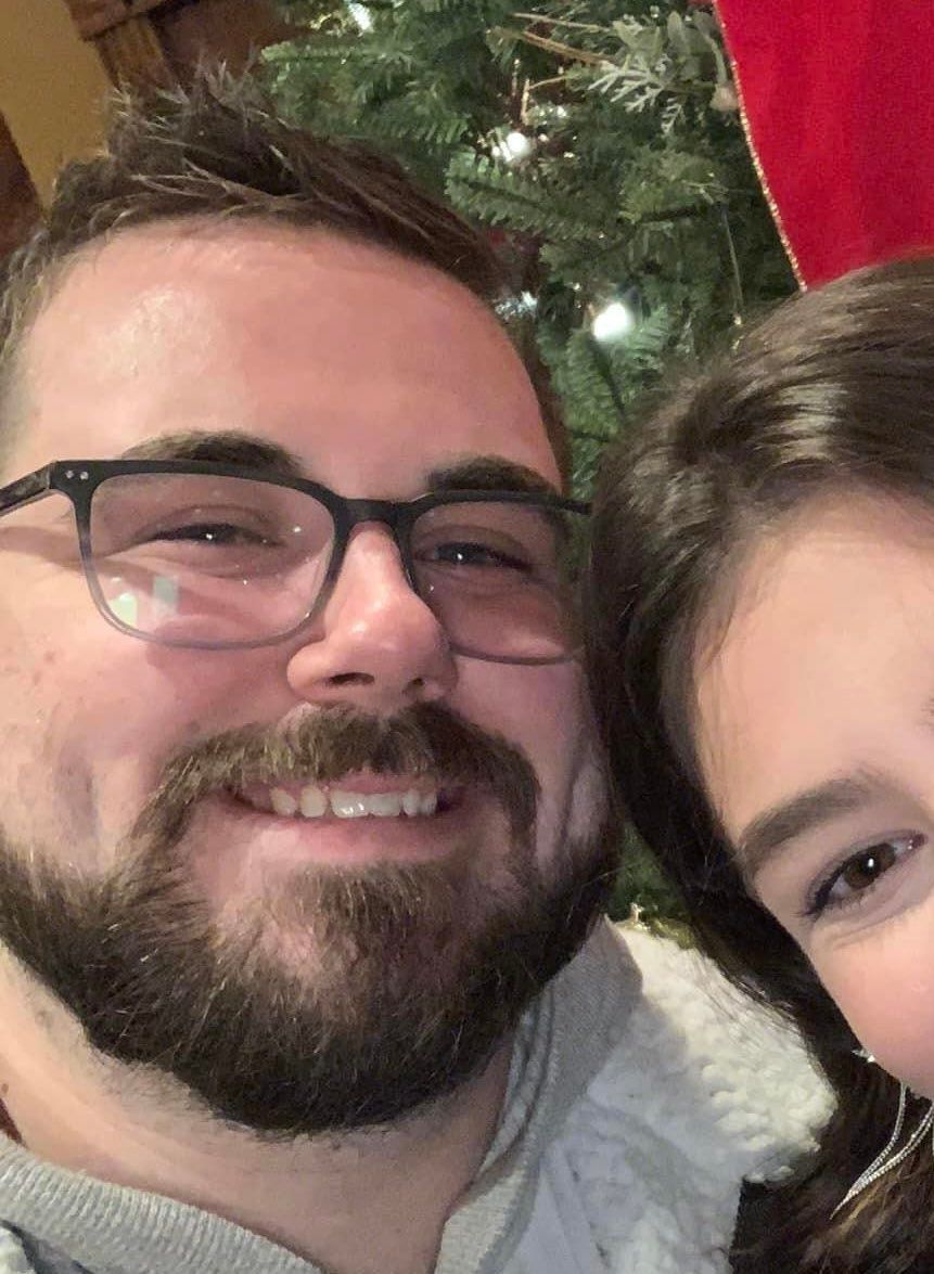 A man with a beard and glasses is smiling next to a woman in front of a christmas tree.