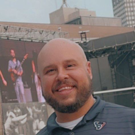 A bald man with a beard is smiling in front of a large screen.