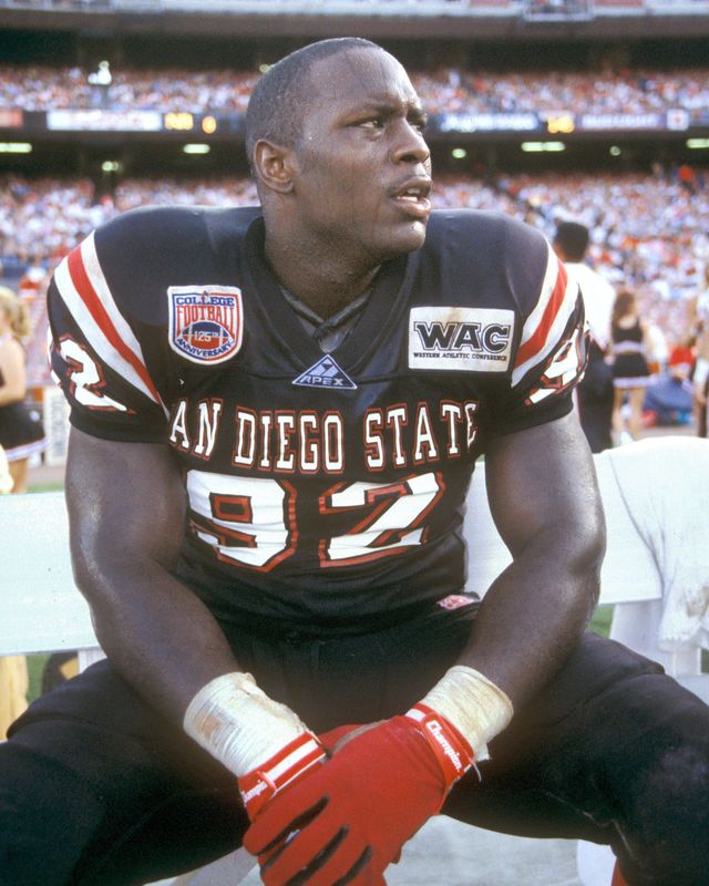 A man in a san diego state 92 jersey sits on a bench