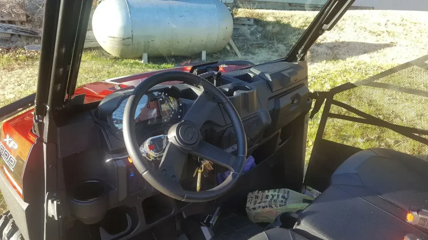 The inside of a utility vehicle with a steering wheel and a propane tank in the background.
