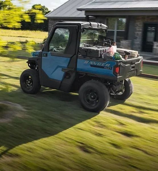 A blue atv is driving down a grassy road in front of a house.