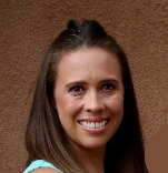 A woman with long brown hair is smiling in front of a brown wall.