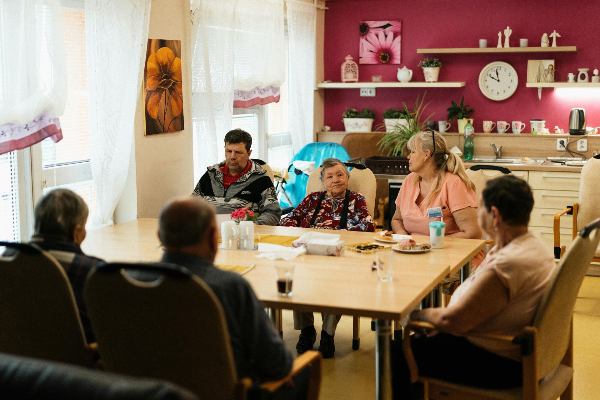 People sit around a dining table in a bright, community-style room with a red accent wall, sharing a social moment.
