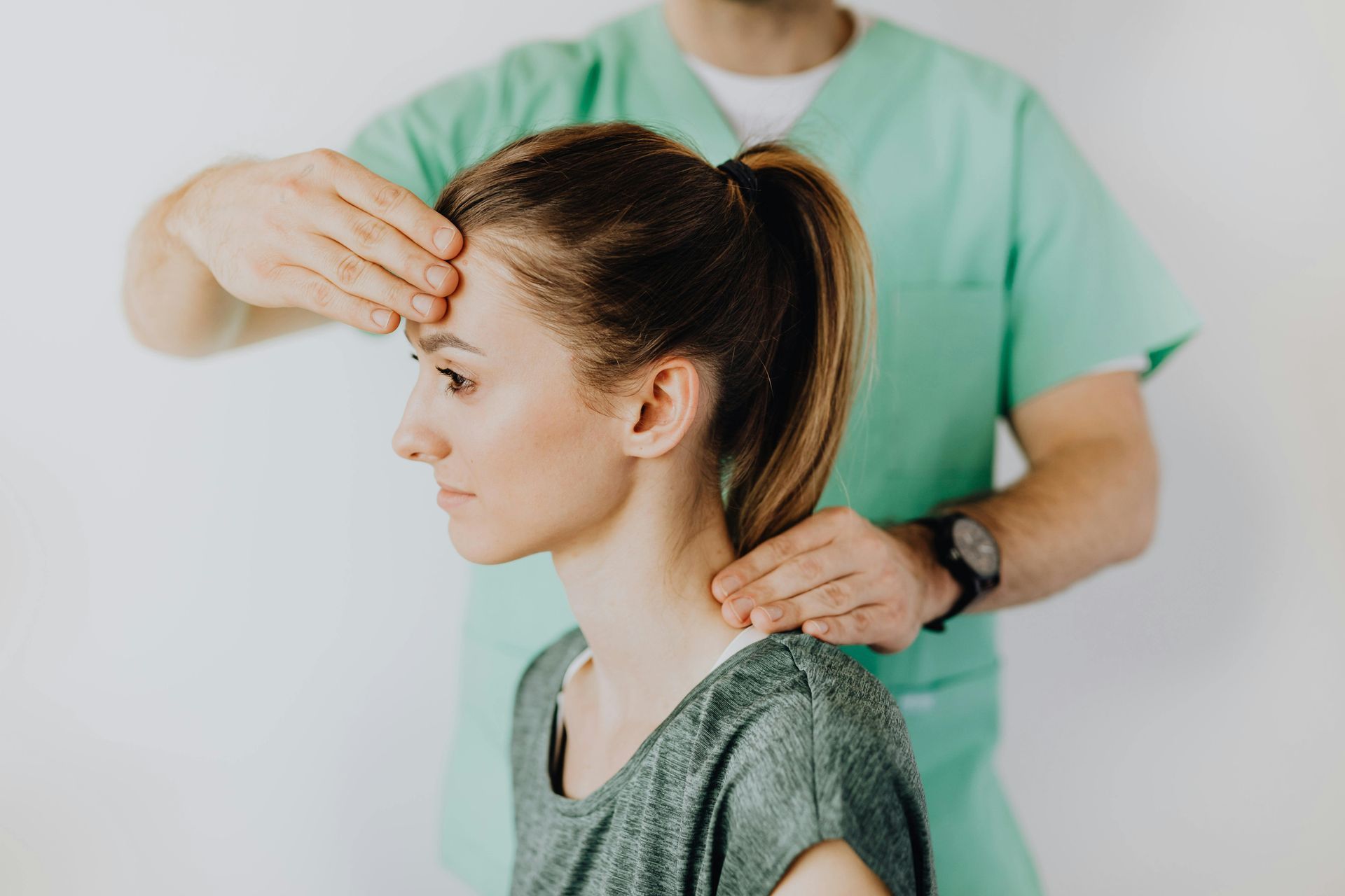 A clinician in green scrubs performs a physical examination by touching a patient’s forehead and the side of their neck.