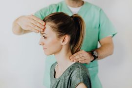 A clinician in green scrubs performs a physical examination by touching a patient’s forehead and the side of their neck.
