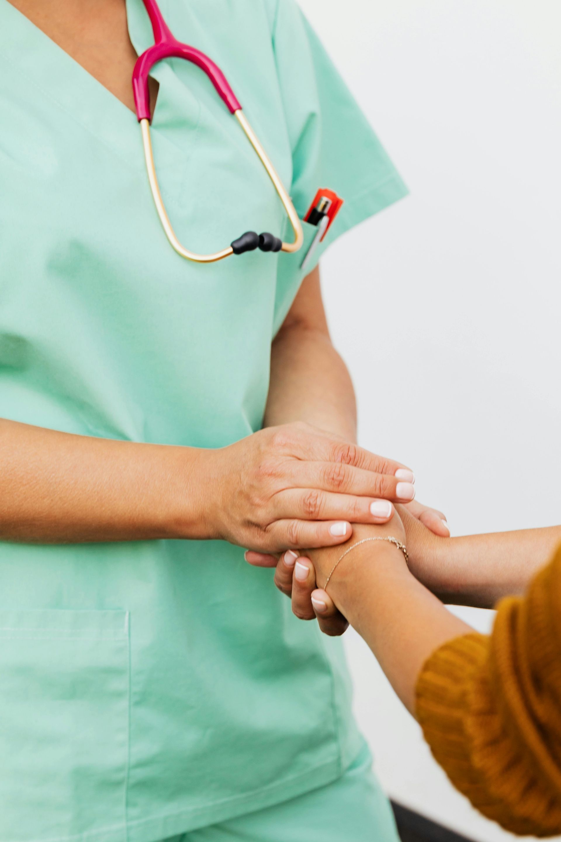 A healthcare worker in green scrubs holds a person's hands in a comforting gesture, with a stethoscope around their neck.