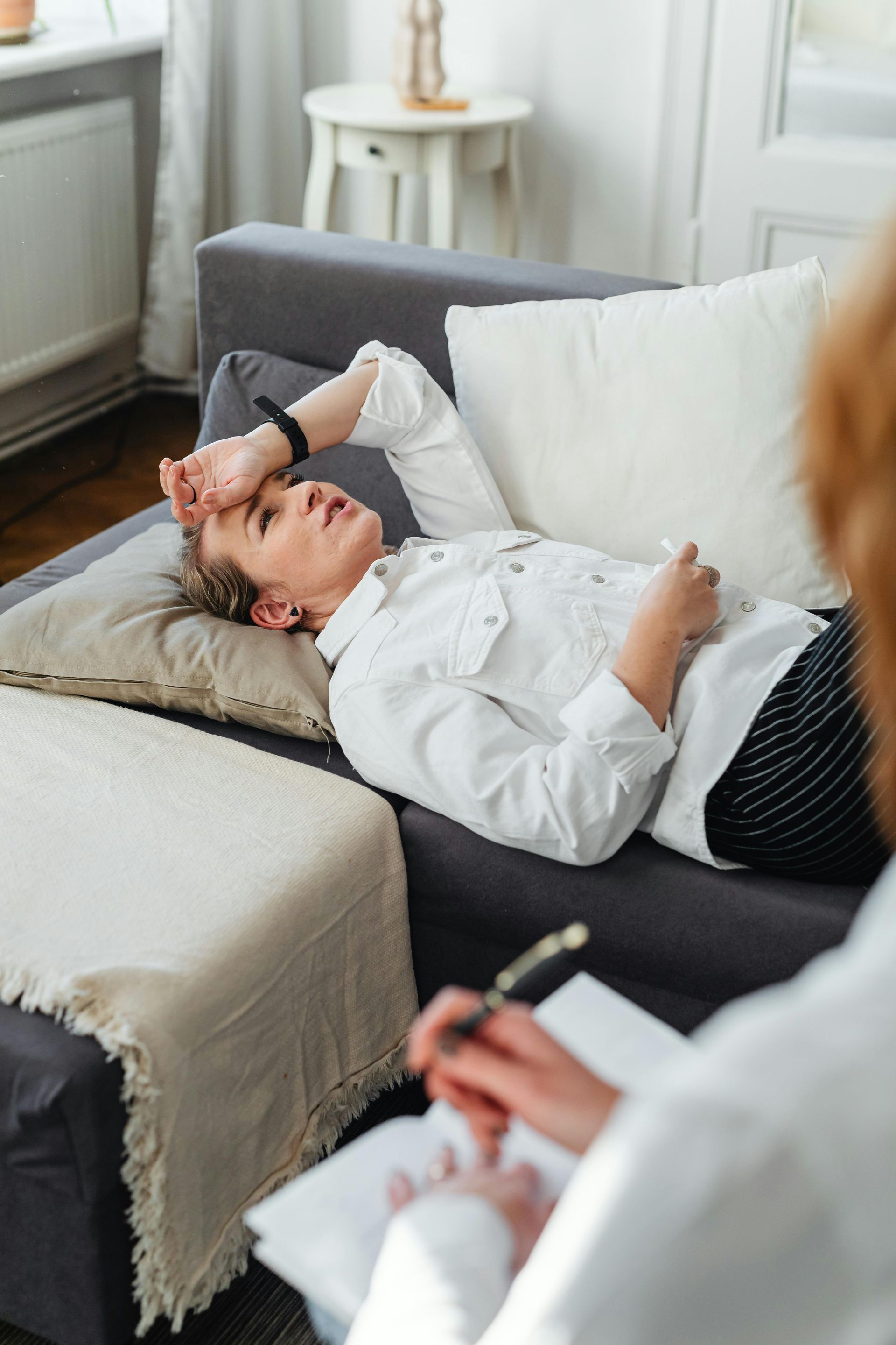 A person lies on a gray sofa with a hand on their forehead, appearing distressed, while a therapist takes notes nearby.