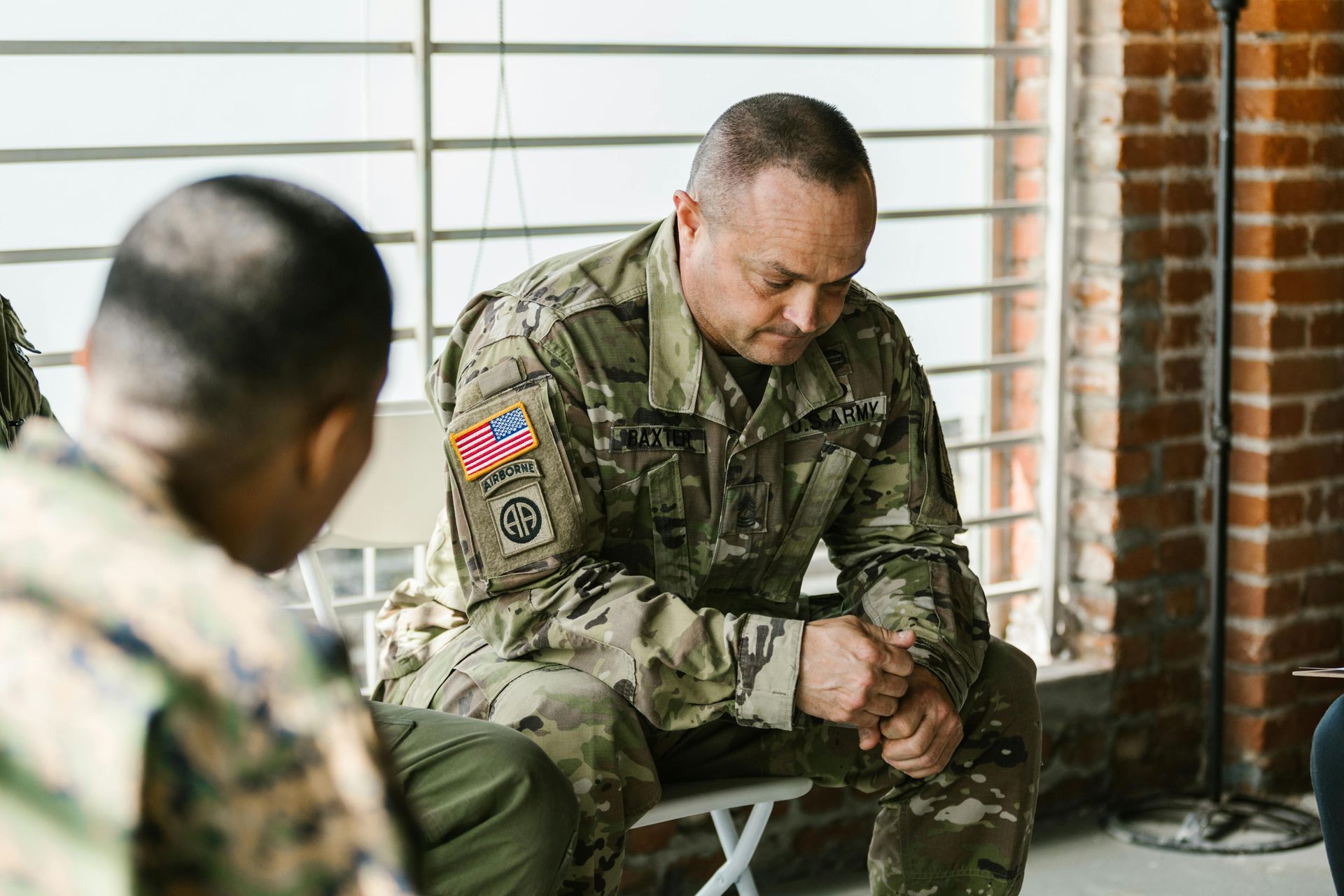 A military member in uniform sitting in a chair, looking downward with a pensive expression during a group meeting.
