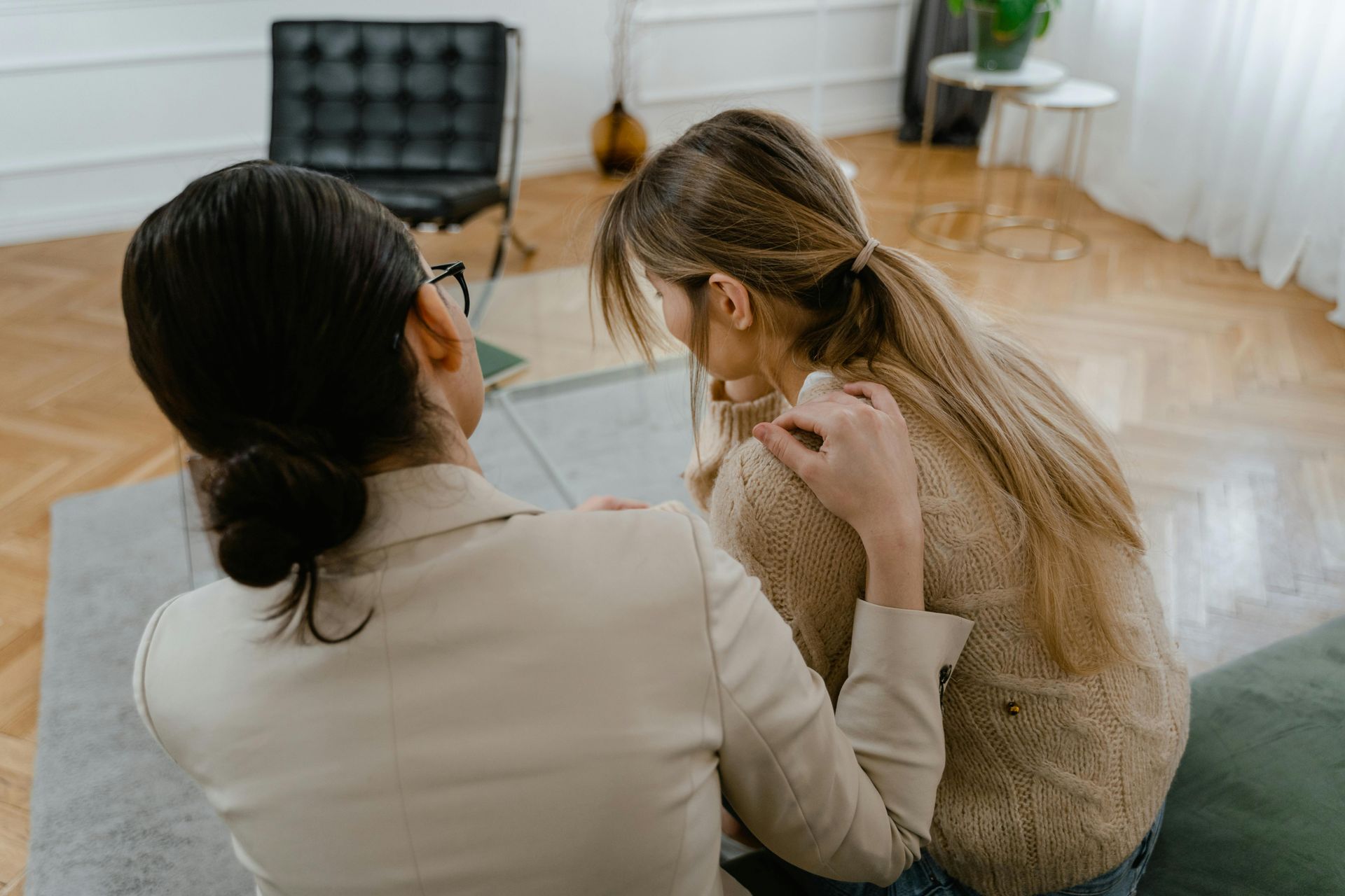 A person with dark hair in a beige blazer comforts a long-haired person sitting on a sofa in a bright room.