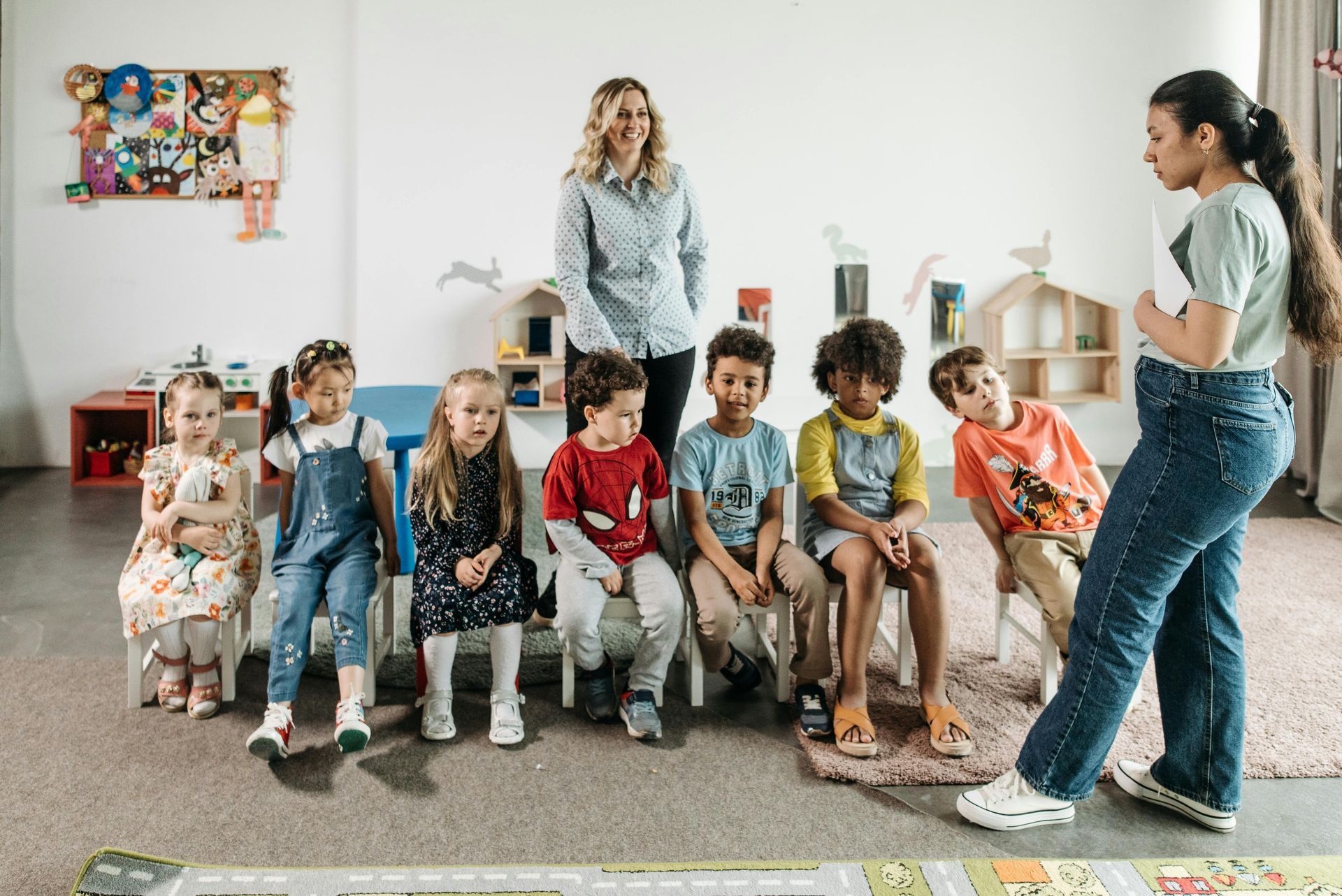 A teacher stands before a group of children seated in a row on chairs in a brightly lit, decorated classroom.