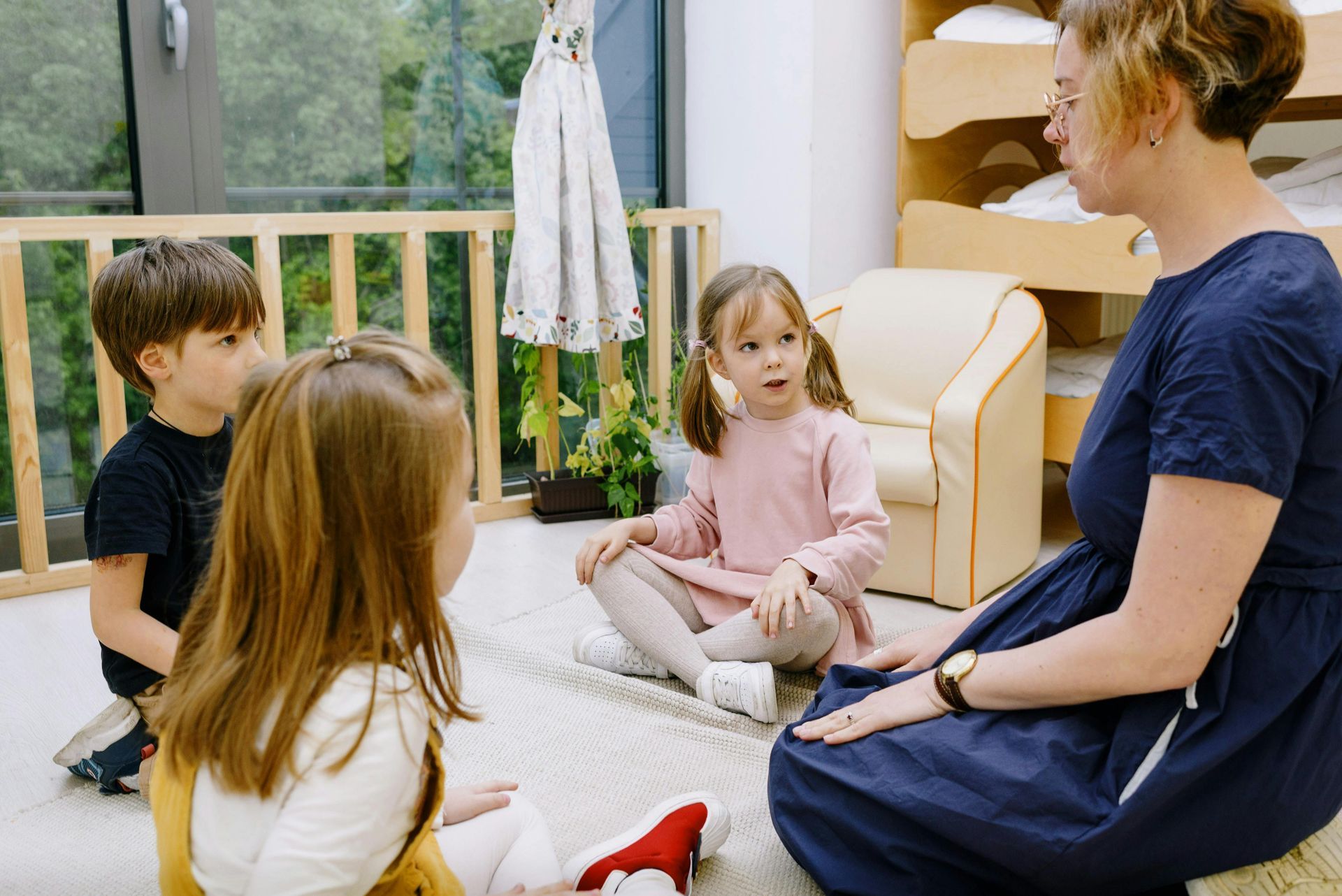 A teacher sits on the floor in a classroom, talking to three children who are gathered around in a circle.