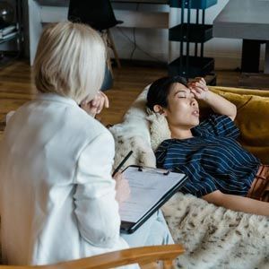 A therapist in a white blazer sits with a clipboard, listening as a person rests on a yellow couch with a hand to their head.