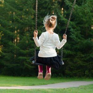 A person with hair tied in a bun, wearing a white shirt and dark skirt, swings on a rope swing before a forest background.