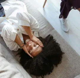 A person with dark curly hair lies on a textured couch, resting their face in their hands, near someone in white sneakers.