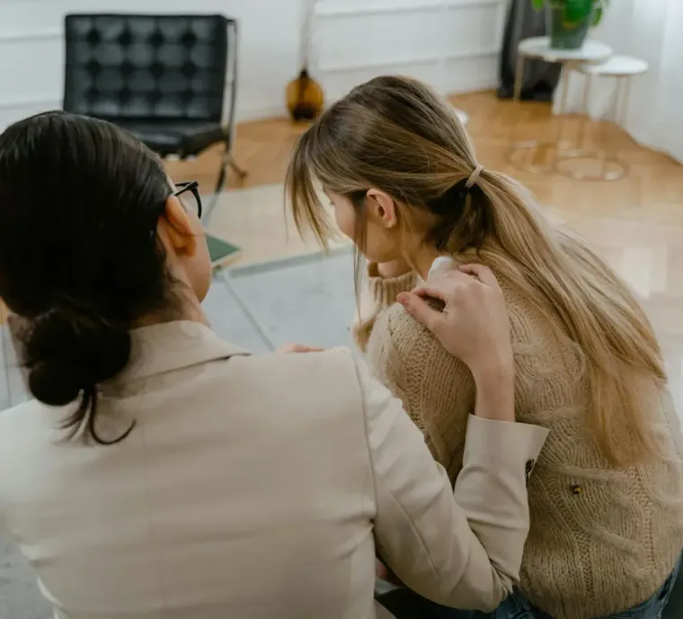 A person with dark hair in a beige blazer comforts a long-haired person sitting on a sofa in a bright room.