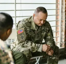 A military member in uniform sitting in a chair, looking downward with a pensive expression during a group meeting.