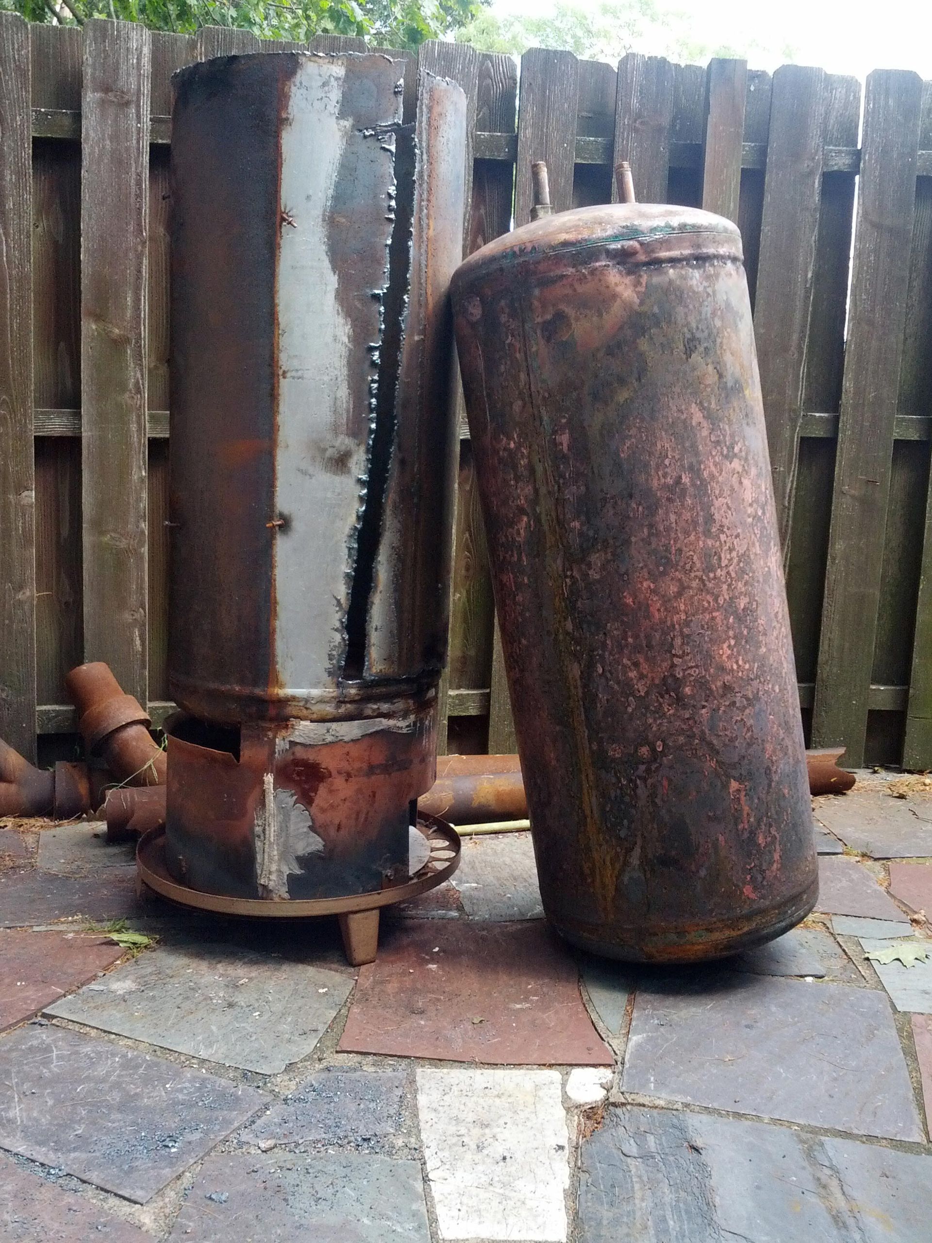 Two rusty metal cylinders are sitting on a sidewalk in front of a wooden fence.
