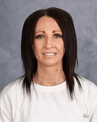 A woman with long brown hair is wearing a white shirt and smiling for the camera.